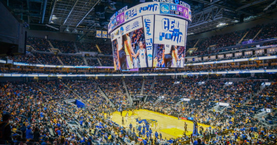 图片展示了一场室内篮球比赛，球场中心有正在比赛的球员，看台上观众众多，大屏幕显示着比赛信息和球员画面。